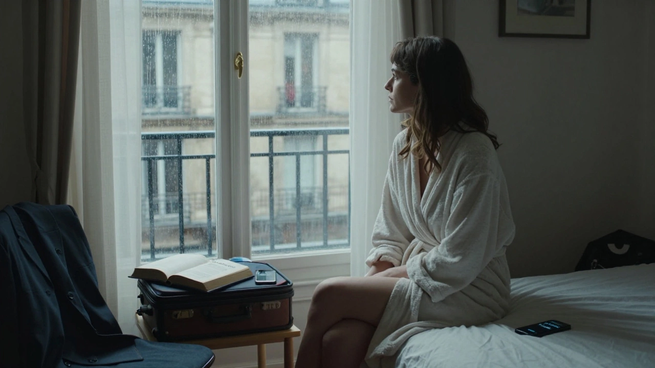 A woman sits by a window in her modest Paris room, textbooks and suitcase nearby, gazing at the rain.